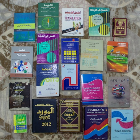 A collection of various books related to translation and dictionaries, with covers in a mix of English and Arabic languages. The books have diverse cover designs featuring text, globes, and other graphical elements. They're arranged in neat rows on a patterned surface.