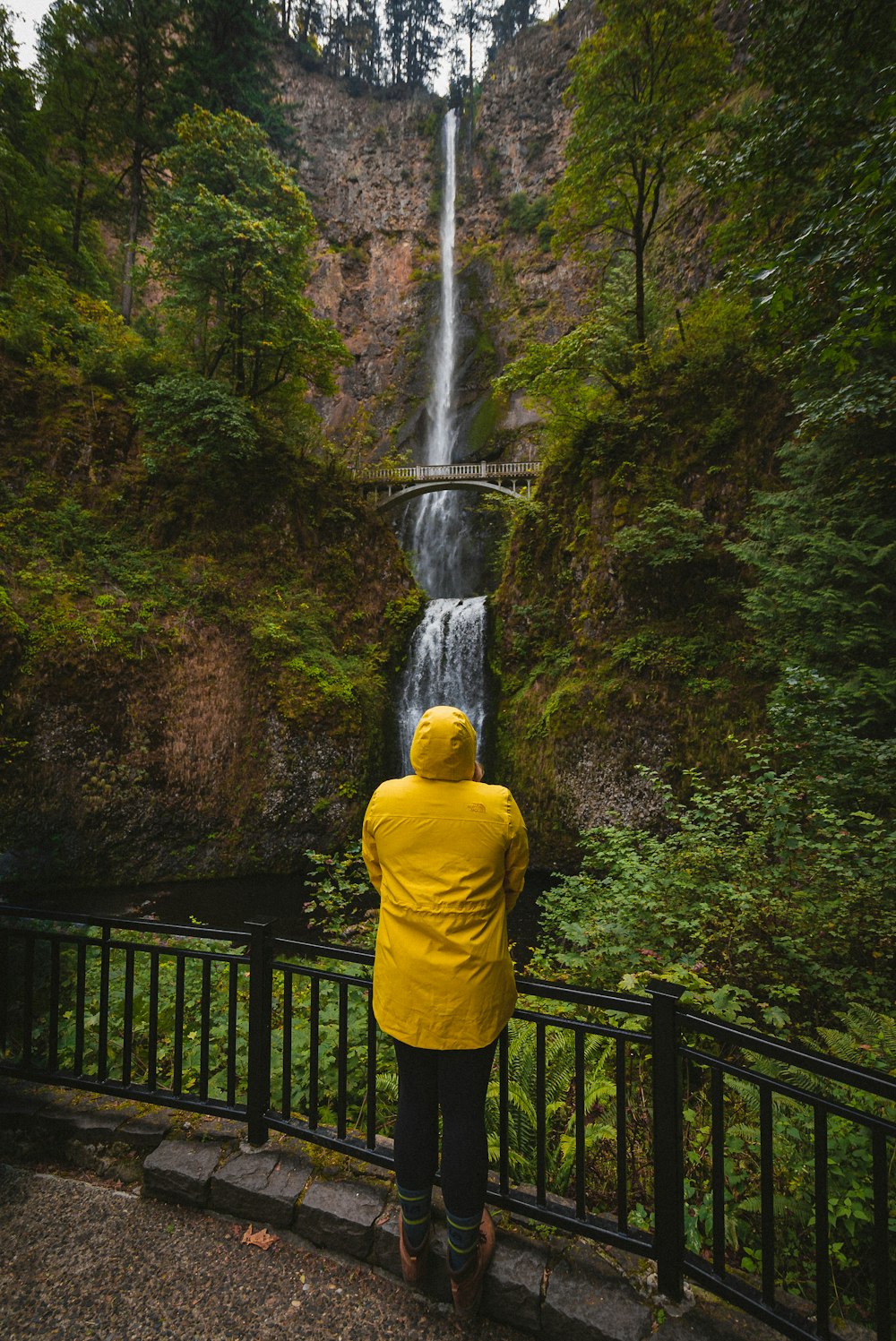 Person in gelbem Kapuzenpulli, die tagsüber auf einer schwarzen Metallbrücke in der Nähe von Wasserfällen steht