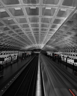 A modern underground subway station with a futuristic design is captured. The ceiling is made up of geometric, concrete patterns. The image features a train speeding through the station, creating a blurred effect that signifies motion. Sparse lighting and passengers can be seen on the platform along the sides.