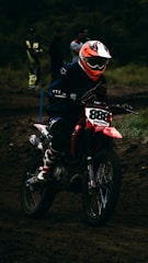 A motocross rider wearing a helmet and protective gear is maneuvering a red dirt bike on a muddy track. The background shows several spectators, some dressed casually and others in racing gear, watching the event.
