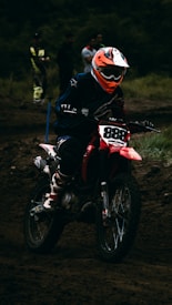 A motocross rider wearing a helmet and protective gear is maneuvering a red dirt bike on a muddy track. The background shows several spectators, some dressed casually and others in racing gear, watching the event.