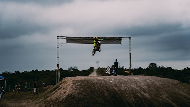 Rider jumping over a dirt ramp on an off-road circuit under a clear sky.