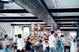 A spacious, modern cafe filled with people, some seated and others standing around. The interior features a mix of industrial and natural decor, including plants and wooden elements. Soft natural light filters in through large windows, creating a warm atmosphere.