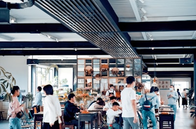 A spacious, modern cafe filled with people, some seated and others standing around. The interior features a mix of industrial and natural decor, including plants and wooden elements. Soft natural light filters in through large windows, creating a warm atmosphere.