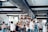 A spacious, modern cafe filled with people, some seated and others standing around. The interior features a mix of industrial and natural decor, including plants and wooden elements. Soft natural light filters in through large windows, creating a warm atmosphere.