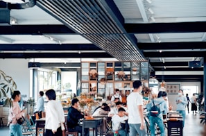 A spacious, modern cafe filled with people, some seated and others standing around. The interior features a mix of industrial and natural decor, including plants and wooden elements. Soft natural light filters in through large windows, creating a warm atmosphere.