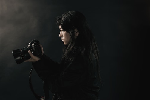 A person with long dark hair holds a DSLR camera up close to their face, appearing focused. The background is dark, creating a moody atmosphere, with subtle lighting highlighting the person's profile and the camera.