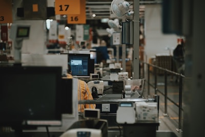 A busy checkout area in a store, featuring several checkout registers each equipped with monitors and scanners. Above the registers, there are signs with numbers. A worker in a striped shirt is visible behind one of the monitors. Various small objects and fans are placed around the area to provide convenience for staff.
