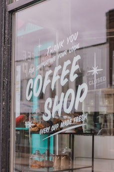 A large glass window displays the words 'Thank you for supporting your local coffee shop', followed by 'Every cup made fresh'. Behind the glass, there are glass jars filled with pastries and two paper bags, presumably holding coffee beans or similar items.