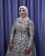 A person is dressed in an elegant, long-sleeved gown with intricate silver and black patterns. The individual is wearing a beige hijab and stands in front of a purple curtain backdrop. The person is smiling, with their hand adorned with henna designs and gold bracelets.
