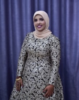 A person is dressed in an elegant, long-sleeved gown with intricate silver and black patterns. The individual is wearing a beige hijab and stands in front of a purple curtain backdrop. The person is smiling, with their hand adorned with henna designs and gold bracelets.