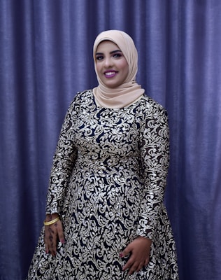 A person is dressed in an elegant, long-sleeved gown with intricate silver and black patterns. The individual is wearing a beige hijab and stands in front of a purple curtain backdrop. The person is smiling, with their hand adorned with henna designs and gold bracelets.