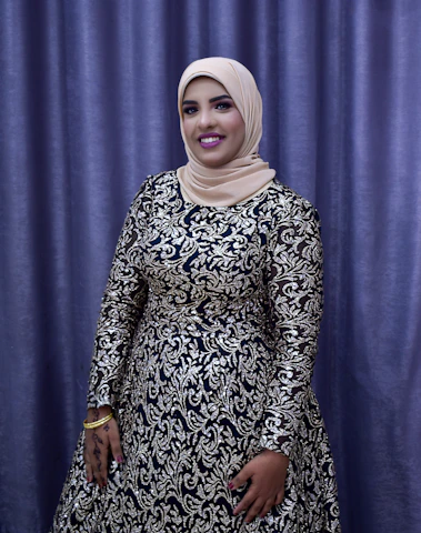 A person is dressed in an elegant, long-sleeved gown with intricate silver and black patterns. The individual is wearing a beige hijab and stands in front of a purple curtain backdrop. The person is smiling, with their hand adorned with henna designs and gold bracelets.