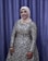 A person is dressed in an elegant, long-sleeved gown with intricate silver and black patterns. The individual is wearing a beige hijab and stands in front of a purple curtain backdrop. The person is smiling, with their hand adorned with henna designs and gold bracelets.