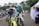 Two motorcyclists in racing gear are riding dirt bikes along a paved path. Several scooters are parked in the background near white tents and palm trees. A man in a blue and white outfit is standing by a tent on the right.