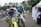 Two motorcyclists in racing gear are riding dirt bikes along a paved path. Several scooters are parked in the background near white tents and palm trees. A man in a blue and white outfit is standing by a tent on the right.