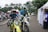 Two motorcyclists in racing gear are riding dirt bikes along a paved path. Several scooters are parked in the background near white tents and palm trees. A man in a blue and white outfit is standing by a tent on the right.