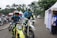 Two motorcyclists in racing gear are riding dirt bikes along a paved path. Several scooters are parked in the background near white tents and palm trees. A man in a blue and white outfit is standing by a tent on the right.
