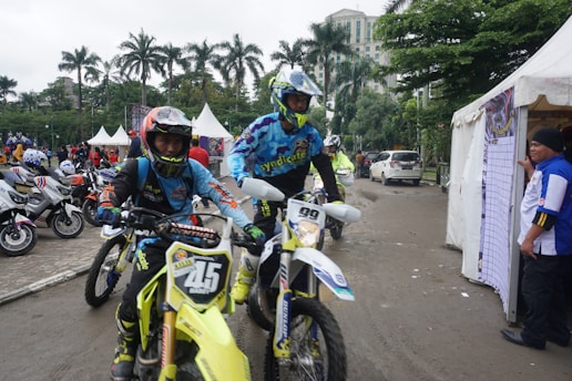Two motorcyclists in racing gear are riding dirt bikes along a paved path. Several scooters are parked in the background near white tents and palm trees. A man in a blue and white outfit is standing by a tent on the right.