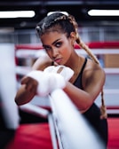 A woman with braided hair is intensely focused, wrapping her hands with boxing tape inside a boxing ring. She has an athletic build and is wearing workout attire. The background features blurred red and white ropes typically found in a gym setting.