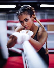 A woman with braided hair is intensely focused, wrapping her hands with boxing tape inside a boxing ring. She has an athletic build and is wearing workout attire. The background features blurred red and white ropes typically found in a gym setting.