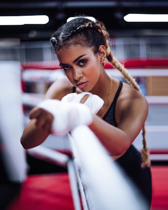 A woman with braided hair is intensely focused, wrapping her hands with boxing tape inside a boxing ring. She has an athletic build and is wearing workout attire. The background features blurred red and white ropes typically found in a gym setting.