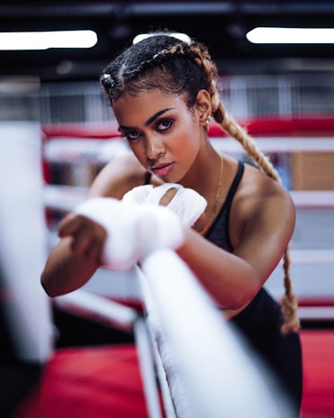 A woman with braided hair is intensely focused, wrapping her hands with boxing tape inside a boxing ring. She has an athletic build and is wearing workout attire. The background features blurred red and white ropes typically found in a gym setting.