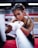 A woman with braided hair is intensely focused, wrapping her hands with boxing tape inside a boxing ring. She has an athletic build and is wearing workout attire. The background features blurred red and white ropes typically found in a gym setting.