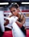 A woman with braided hair is intensely focused, wrapping her hands with boxing tape inside a boxing ring. She has an athletic build and is wearing workout attire. The background features blurred red and white ropes typically found in a gym setting.