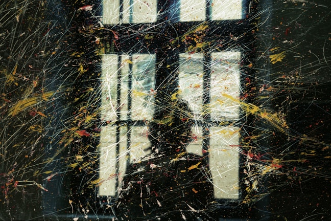 A window is seen with a reflection that appears to be partially obscured by various streaks and splatters of paint. The colors are scattered across the surface, creating an abstract and dynamic visual pattern.