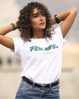 A person with curly hair wearing a white T-shirt that has the word 'FEELINGS' printed on it. They have their hands resting on their head and are wearing blue jeans with a black belt featuring a gold logo. The background is blurred, suggesting an outdoor setting.