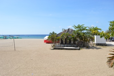 A sandy beach stretches towards the blue ocean, with a small hut surrounded by palm trees positioned near the center. In the background, a few people can be seen swimming, while boats are visible on the water. A volleyball net is set up on the left side, and white and red tents are to the right. The sky is clear and blue, enhancing the tropical feel.