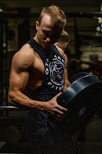 A muscular man is lifting a heavy barbell plate in a darkly lit gym. He is wearing a sleeveless gym shirt that accentuates his defined biceps and triceps. The dim lighting creates strong contrasts and highlights his muscular definition.