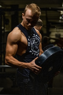 A muscular man is lifting a heavy barbell plate in a darkly lit gym. He is wearing a sleeveless gym shirt that accentuates his defined biceps and triceps. The dim lighting creates strong contrasts and highlights his muscular definition.