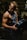 A muscular man is lifting a heavy barbell plate in a darkly lit gym. He is wearing a sleeveless gym shirt that accentuates his defined biceps and triceps. The dim lighting creates strong contrasts and highlights his muscular definition.
