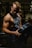 A muscular man is lifting a heavy barbell plate in a darkly lit gym. He is wearing a sleeveless gym shirt that accentuates his defined biceps and triceps. The dim lighting creates strong contrasts and highlights his muscular definition.