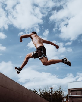 A person is dynamically jumping across a gap against a background of a bright blue sky with scattered white clouds. The individual is wearing athletic shorts and shoes, displaying strong movement and athleticism.