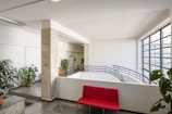 A bright indoor space featuring large windows with metal frames that allow natural light to fill the area. There are multiple green potted plants placed around the room, contributing to a fresh and inviting atmosphere. A bold red bench seat stands out against the neutral tones of the walls and floor. The design elements include marble tiles, metal railings, and minimalist decor.