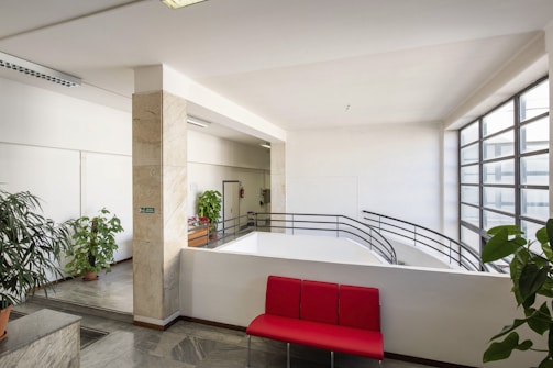 A bright indoor space featuring large windows with metal frames that allow natural light to fill the area. There are multiple green potted plants placed around the room, contributing to a fresh and inviting atmosphere. A bold red bench seat stands out against the neutral tones of the walls and floor. The design elements include marble tiles, metal railings, and minimalist decor.