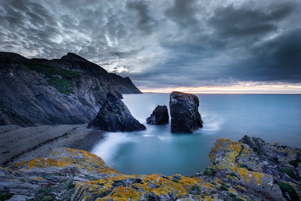 brown and green rock formation near body of water under gray clouds