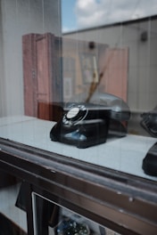 A black rotary dial telephone is displayed behind a glass window. The reflection of the outside is visible on the glass surface. The background shows a blurred setting with a few wooden furniture pieces.