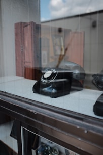 A black rotary dial telephone is displayed behind a glass window. The reflection of the outside is visible on the glass surface. The background shows a blurred setting with a few wooden furniture pieces.