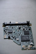 A close-up view of a computer circuit board with numerous integrated circuits, resistors, and capacitors visible. The board has a variety of small components scattered across its surface, including microchips labeled SMOOTH and TDK. The background is a neutral, smooth surface.