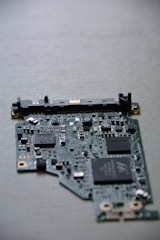 A close-up view of a computer circuit board with numerous integrated circuits, resistors, and capacitors visible. The board has a variety of small components scattered across its surface, including microchips labeled SMOOTH and TDK. The background is a neutral, smooth surface.