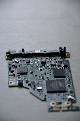 A close-up view of a computer circuit board with numerous integrated circuits, resistors, and capacitors visible. The board has a variety of small components scattered across its surface, including microchips labeled SMOOTH and TDK. The background is a neutral, smooth surface.