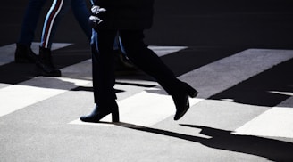 People are crossing a street at a pedestrian crosswalk. The scene includes several individuals walking, with strong shadows cast by bright sunlight. The focus is on the legs and lower bodies of the pedestrians, emphasizing their movement.