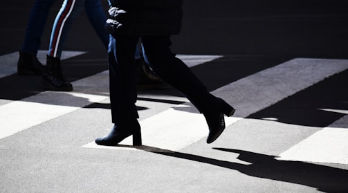 People are crossing a street at a pedestrian crosswalk. The scene includes several individuals walking, with strong shadows cast by bright sunlight. The focus is on the legs and lower bodies of the pedestrians, emphasizing their movement.