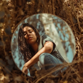 A woman's face is reflected in a round mirror placed among dry brown foliage. Her expression is calm, and she is wearing a casual greenish-blue shirt. Her dark hair is slightly tousled, and the background features muted tones, creating an earthy, natural atmosphere.