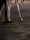 A person is standing on a paved surface, wearing grey pants and brown shoes. They are holding a large brush, which they use to write on the ground. The brush is attached to a long handle, and their writing appears to be artistic in nature. Others are walking in the background.