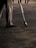 A person is standing on a paved surface, wearing grey pants and brown shoes. They are holding a large brush, which they use to write on the ground. The brush is attached to a long handle, and their writing appears to be artistic in nature. Others are walking in the background.